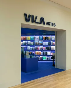Entrance view of a modern bookstore with intense blue interior lighting, bookshelves, and a blue display platform, featuring a dark blue 'VILA ARTES' sign above the doorway.