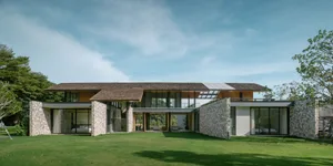 Wide exterior view of a modern stone and wood courtyard villa with large glass openings, set in a lush green lawn under a blue sky.