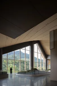 Spacious modern lobby interior with polished concrete floor, curved wooden ceiling, massive floor-to-ceiling windows overlooking green mountains, and a person standing near the glass.