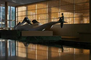 Modern abstract white marble sculpture with a bronze seated figure displayed on a green marble platform in a sunlit contemporary lobby with woven wooden lattice screens.