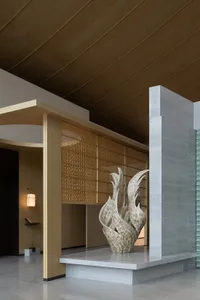 Modern minimalist entrance hall featuring a large abstract travertine flame sculpture on a stone pedestal, with wood lattice screen, concrete pillar, and warm wood ceiling.