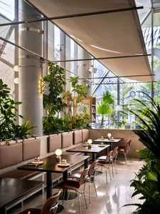 Modern cafe interior featuring large concrete pillars, built-in upholstered banquettes, wooden tables, brown leather chairs, and numerous potted plants under a high fabric canopy with natural daylight streaming through full-height windows.