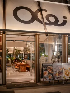 Night view of Ocoi restaurant entrance with open glass doors revealing a long warm wooden communal table, lush indoor plants, and elegant modern lighting.