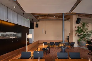 Modern industrial-style restaurant interior with wooden tables, dark blue chairs, built-in tan banquette seating, a central cylindrical steel fireplace, and warm ambient lighting.
