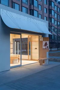 Modern minimalist boutique storefront at dusk with illuminated oak display shelves, concrete bench, and large glass entrance.