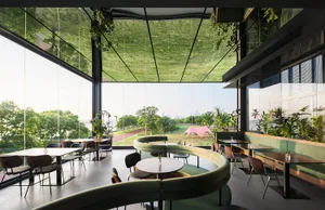 Modern restaurant interior featuring a large curved green velvet booth, wooden tables, black chairs, abundant indoor plants, and floor-to-ceiling glass walls overlooking a riverside park and city skyline.