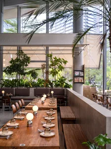Modern biophilic restaurant interior featuring long walnut communal tables, wooden benches, spherical pendant lamps, abundant indoor plants, and large windows with city views.
