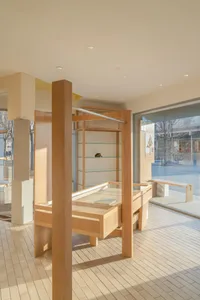 Modern minimalist boutique interior featuring a custom maple wood display fixture with glass shelves and integrated drawers, bathed in warm natural light from large windows.