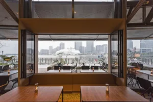 Modern restaurant interior featuring wooden tables, built-in seating, and large foldable glass doors opening to a waterfront city view with distinctive white architecture in the distance.