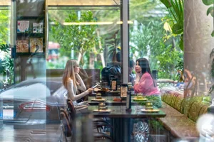 Two women sitting at a table inside a modern glass-walled restaurant overlooking a lush green garden, with marble bench seating and warm wooden tones.