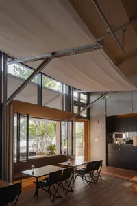 Modern cafe interior featuring a large retractable beige fabric awning, corten steel details, wooden tables, black folding chairs, and full-height glass doors opening to an outdoor terrace.