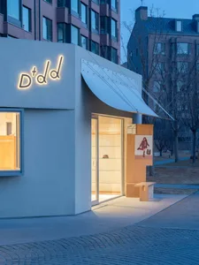 Modern minimalist retail storefront with concrete facade, glowing neon-style logo, large glass entrance, and wooden display elements at dusk.