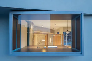 View through a large blue steel window frame into a minimalist boutique store interior featuring oak wood cabinetry, concrete walls, and a dark display table with a modern lamp.