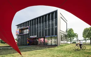 Modern two-story commercial building with dark perforated metal facade and colorful graphic branding for Giant and Napan, set beside a lake with a cyclist passing in the foreground.