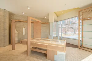 Modern minimalist boutique interior with a large light oak central display island featuring integrated glass display cases, matching wooden clothing racks against a beige brick wall, and large windows providing natural light.