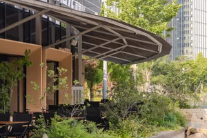 Modern restaurant outdoor seating area featuring a large curved corten steel canopy, black furniture, and abundant greenery with modern buildings in the background.