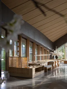 Modern Japanese-style dining hall featuring a long light oak communal table, woven rattan partition, concrete walls, wooden ceiling, and large windows overlooking greenery.