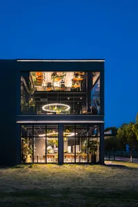 Night view of a modern two-story glass cube house with fully illuminated interior, showing dining area, plants, and circular light fixtures against a dark blue sky.