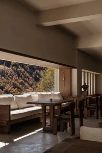 Modern alpine dining room featuring a built-in walnut banquette with cream linen cushions, dark walnut dining table and chairs, concrete floor, and a large panoramic window overlooking an autumnal mountain forest.