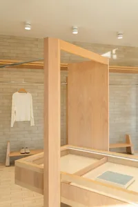 Modern minimalist retail interior with light oak wood display counters, shelving systems, and beige brick walls, featuring a white long-sleeve shirt hanging on a clothing rack and a jewelry display tray.