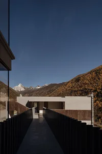 Modern minimalist concrete house with wooden walkway and railing overlooking snow-capped mountains and autumn-colored forest under clear blue sky.