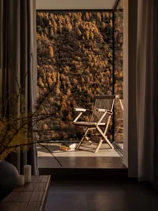 Minimalist interior with an oak wooden folding chair placed by a floor-to-ceiling window overlooking an autumn mountain forest at golden hour