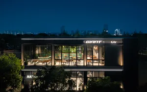 Night view of a modern two-story glass facade restaurant with illuminated interior seating, curved green banquettes, and lush indoor plants, branded as Giant Liv.