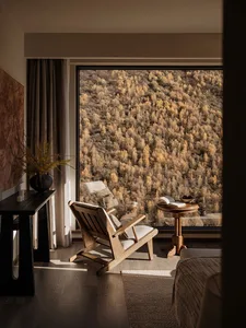 Wooden reading chair and side table placed beside a large picture window overlooking a vast autumn mountain forest in warm golden light.