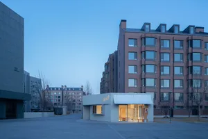 現代極簡單層混凝土零售建築，搭配白色遮陽棚與明亮室內照明，位於黃昏時分的都市廣場，背景為磚造公寓大樓。