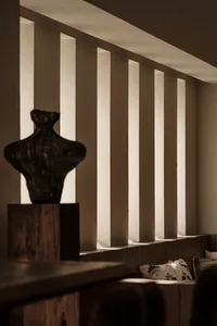 Interior view of a modern living space featuring a textured off-white wall with vertical light strips, dark wood paneling, a wooden pedestal with an abstract dark sculpture, and patterned cushions on a built-in bench.