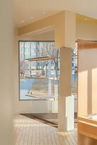Modern minimalist entryway featuring a travertine stone pillar, three floating wooden shelves behind glass, light wood cabinetry, and large windows overlooking a courtyard with bare trees.