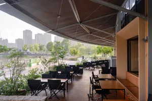 Modern outdoor restaurant patio featuring wooden decking, black folding chairs and tables, under a large fabric canopy with lush greenery and a river view in the background.