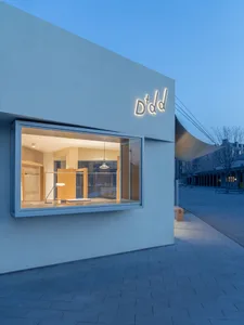 Exterior view of a modern minimalist concrete building with a large illuminated display window showing warm wooden interior elements and a hanging pendant lamp at dusk.