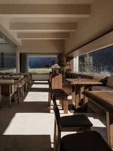 Modern rustic dining room interior with solid wood tables and chairs, concrete floor, large windows overlooking mountains and grassland.
