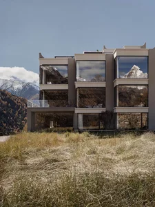 Modern multi-story concrete and glass mountain house with large panoramic windows reflecting snow-capped alpine peaks, set against a foreground of wild mountain grass under a clear blue sky.