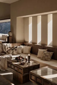 Modern minimalist living room featuring a built-in walnut wood banquette seating with cream cushions, patterned throw pillows, and dark wooden coffee tables under dramatic natural light from vertical wall slots.