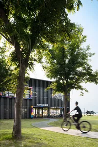 Modern dark corrugated metal cultural building with colorful window accents, surrounded by large trees and a winding bicycle path, with a cyclist in motion in the foreground.