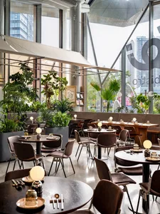 Modern restaurant interior featuring round wooden tables, brown upholstered chairs, numerous indoor plants, large floor-to-ceiling windows with city views, and spherical pendant lights.