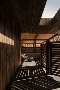 A modern sunken outdoor patio entirely clad in dark wood with dramatic diagonal sunlight shadows, featuring a rattan lounge chair, stone coffee table, and mountain forest view in the background.