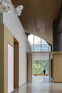 Modern minimalist entrance hall featuring a sweeping curved wooden ceiling, polished concrete floor, large floor-to-ceiling windows overlooking green mountains, and a person standing in the distance.
