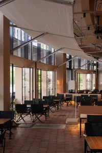 Modern restaurant interior featuring terracotta floor tiles, black folding chairs, wooden tables, large glass windows, and large suspended white canvas canopies creating a warm industrial atmosphere.