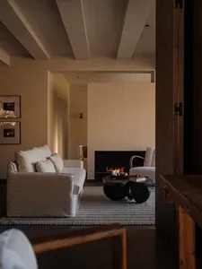Modern minimalist living room featuring a white linen sofa, sculptural black coffee table, lit fireplace, and textured beige plaster walls with exposed wooden ceiling beams.