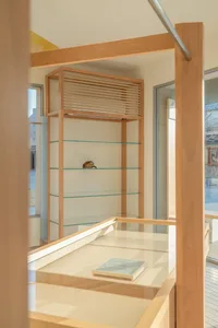 Modern minimalist oak wood jewelry display cabinet featuring tempered glass shelves and a glass-top counter, with a baseball cap and pearl necklace on display.