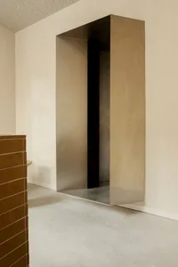 Modern minimalist interior featuring a large brushed stainless steel pivot door with dark reveal, set against off-white walls and concrete floor, with a terracotta brick element in the foreground.