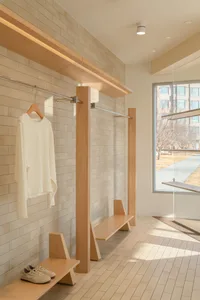 Modern minimalist mudroom featuring light oak floating benches, white subway tile walls, chrome clothing rack, and large window with natural sunlight.