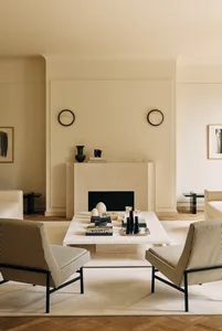 Modern minimalist living room featuring a large travertine fireplace, two striped upholstered armchairs, a matching travertine coffee table, and neutral beige walls with decorative objects.