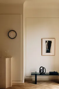 Minimalist entryway featuring a travertine pedestal, round black-framed mirror, black console table, abstract metal sculpture, and framed artwork under warm ambient lighting.