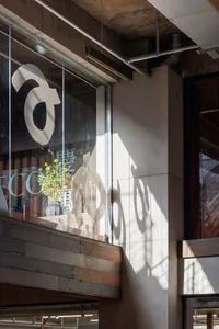 Interior corner of an industrial-style cafe featuring raw concrete walls, reclaimed wood paneling, large glass windows with white Chinese characters, a vase of yellow flowers, and exposed ceiling pipes under bright sunlight.