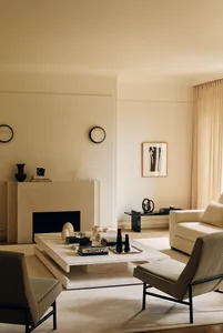 Modern minimalist living room featuring a large travertine coffee table, matching fireplace mantel, two striped armchairs with black metal frames, a cream sofa, and warm ambient lighting.