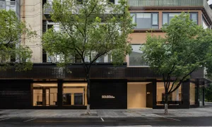 Street view of SOLSO by SOLSO retail store featuring a modern black tiled facade, large glass openings revealing warm-lit interiors, and green trees in front.